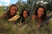 Eva, Laura, and Dani lying in grass looking serious at camera while holding machetes.