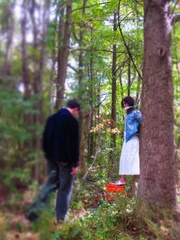 A man looking at a woman who is tied up against a tree in the woods.