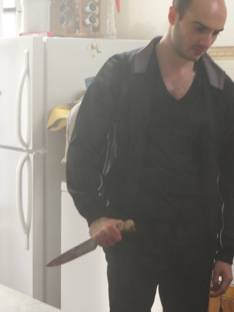 The murderer holding a bloody knife after murdering an older man in the kitchen.