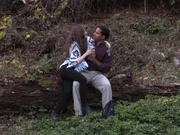 A man and woman sitting on a log in the woods drinking.