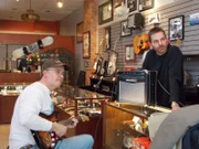 A man playing the guitar in the shop with Scott.