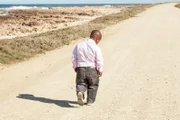 dwarfish african man walking down a dirt road along the coast with his head bent down