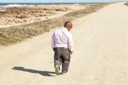 dwarfish african man walking down a dirt road along the coast with his head bent down