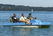 Hubert (Christian Tramitz, Mitte) und Staller (Helmfried von Lüttichau, rechts) sitzen in einem Tretboot und fahren auf den See hinaus. Yazid (Hannes Ringelstetter) sitzt hinter ihnen in Tauchermontur. Er soll für Hubert und Staller testen, ob man mit einem Schlittschuh unter Wasser an einer Boje hängen bleiben kann.