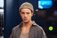 Duncan (Jonas Holdenrieder), mit grauer Mütze, grauem Shirt und einem grauen Hemd darüber, steht in seinem Atelier.