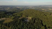 Luftaufnahme eines bewaldeten Berggipfels in hügeliger Landschaft. Auf dem Gipfel sind Umrisse ausgegrabener Gebäude und weiße Zelte zu erkennen.