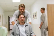 Quirin Pankofer (Jens Atzorn) schiebt Markus Edlinger (Oliver Rosskopf) im Rollstuhl durch den Krankenhausflur, während Lena Lorenz (Patricia Aulitzky) den beiden nachschaut.