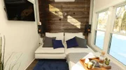 Sterling Folkestad's tiny house features this sleek, modern living room with cozy white couches, a huge flat screen and a cozy bed that sits on a pulley system right above the living room and can be lowered down to be used by visiting friends and family here in Ipswich, Massachusetts, as seen on Tiny House, Big Living.