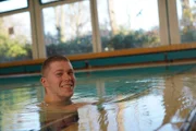Niklas ist im Schwimmbecken, hält sich am Beckenrand fest und lacht.