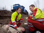 Dr. Elena Eichhorn (Cheryl Shepard, links) trifft mit einem Kollegen (Komparse, rechts) am Unfallort ein. Dr. Ralph Groher (Christoph Grunert, liegend) ist dem Zug zwar im letzten Moment ausgewichen, aber dabei ist er gestürzt und hat sich eine Beinfraktur zugezogen.