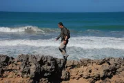 Bear Grylls walking over rocks with waves breaking in the background.