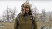 Ed Stafford close-up wearing fur hat with snowy trees and rock in background.