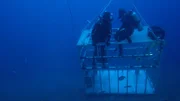 Divers sit on top of cage on ocean floor.