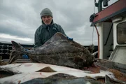 Landscape MS of Jeremy Wade holding a halibut on board of a fishing trawler. Jeremy looking directly at camera against a cloudy sky. Location: Dutch Harbor, AK