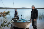 MS landscape of Jeremy Wade standing next to a moored boat on the edge of a lake, Guide Oscar Hedskog standing on the back of the boat next to the engine. Foliage out of focus in foreground.