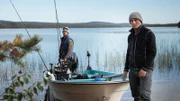 Jeremy Wade standing next to a moored boat on the edge of a lake, Guide Oscar Hedskog standing on the back of the boat next to the engine. Foliage out of focus in foreground.