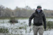 Jeremy Wade wading through shallow reedbeds towards the camera, holding his fishing rod whilst on the right side of the frame.