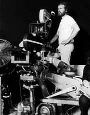 A cameraman stands next to a large camera.