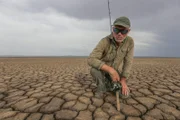 Landscape shot of Jeremy facing towards camera, kneeling with fishing rod on dry cracked river bed Location: Western Australia