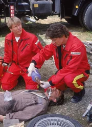 Dr. Mark Harland (Urs Remond, re.) und Peter Berger (Serge Falck, li.) versorgen den schwer verletzten Schrottplatzbesitzer Willmann (Oliver Hebeler, lieg.).