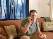 Rob Andrew (Actor, Guy Olivieri) is sitting on the couch while on the phone.