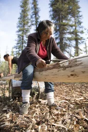 Edna cutting wood while sitting on a log.
