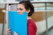 Rebecca (Milena Straube) versucht heimlich etwas zu beobachten und hält sich dazu eine Aktenmappe vors Gesicht.