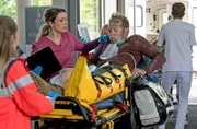 Marco Schiebel (Frederik Schmid, M.) hat bei dem Großunfall eine Fraktur und schwere Verbrennungen erlitten. Viktoria Stadler (Isabella Krieger, 2.v.r.) übernimmt den Patienten in der Notaufnahme von den Sanitäter:innen (Komparsen:innen).