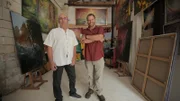 Josh Gates und Michel Becker stehen inmitten der Originalillustrationen für das Golden Owl-Puzzle in Rochefort, Frankreich.