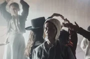 Agnes Sampson standing inside the church amongst the crowd of the witches sabbath. Agnes is staring into to the distance. In the crowd are a few visitors and the devil.