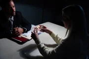 Ein Detektiv beobachtet eine Frau, gegen die ermittelt wird und die ein Bild in der Hand hält.