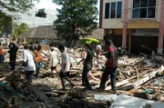 Überlebende laufen durch die Trümmer nach dem Tsunami in Banda Aceh, Indonesien. Ein schweres Erdbeben im Indischen Ozean löst einen Tsunami aus, der innerhalb von Minuten die belebte Stadt Aceh erreicht und eine noch nie dagewesene Verwüstung anrichtet. „Tsunami: Wettlauf gegen die Zeit“ verfolgt den verzweifelten Überlebenskampf von Stadtbewohnern in Indonesien und Touristen in Thailand.