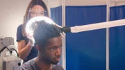 Mid shot of Dr. Emma examining the back of Jelani’s head through lit-up magnifier.