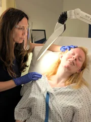 Mid shot of Dr Emma examining Ally’s Skin under lit up magnifier.