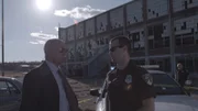 Demery and a cop talk in front a of rundown building.