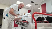 E6 -  Male bald nurse placing an access inlay in the patient's arm. The red handle bars are in the front of the pic, the patient is blurred