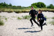 Süher Özlügül (Sophie Dal) und Henk Cassens (Maxim Mehmet) sprinten den Strand entlang zum Wasser. Werden sie es rechtzeitig schaffen?