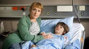 Sonja Kalz (Lotte Ohm, l.) ist bei ihrem Sohn Ole Kalz (Arien Weber, r.), der nach einem Fahrradunfall im Krankenhaus liegt. Ist sie nur deshalb so nervös? Und warum will das Krankenhaus Ole nicht nach Hause lassen, obwohl er dort von seiner Oma Marianne Kalz und Krankenschwestern gepflegt werden könnte?