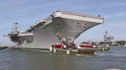 Zwei Spezialschlepper helfen dem 95 tausend Tonnen schweren Flugzeugträger vor Beginn des Manövers aus dem Hafen hinaus auf offene See zu steuern.