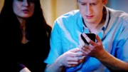 Ein Mann hält ein Mobiltelefon in der Hand, neben ihm sitzt eine Frau.