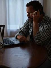Recre of Michael sitting at dining table looking at computer on the phone with hand closest to camera. Side angle shot.