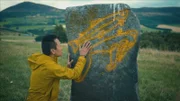 Albert Lin untersucht einen großen stehenden Felsen mit Einritzungen piktischer Symbole, die in diesem computergenerierten Bild grafisch verbessert und hervorgehoben wurden. Tap O'Noth ist im Hintergrund zu sehen.