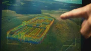 Eine computergenerierte Grafik zeigt Albert Lins Zeigefinger, der auf ein Lidar-Bild einer großen offenen Landschaft zeigt, über das eine CGI einer römischen Siedlung gelegt ist.