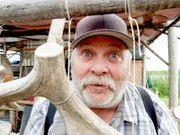 Pat Moore goofing around off set of Yukon Men, Season 3, poking his head through antlers that are hanging at his fish camp.
