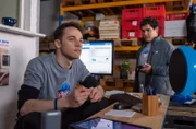 Lenny (Fabian Hanis, l.) sitzt am Empfang von Delivery Ape und spielt gedankenverloren mit dem Stift. Adrian (Danilo Kamperidis, r.) scheint er nicht wahr zu nehmen.