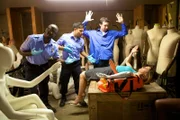 A woman laying on a shipping container in a mannequin room with two EMTs and a man.