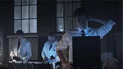 Ein Mann sitzt an einem Schreibtisch. Drei Männer stehen im Kontrollraum.