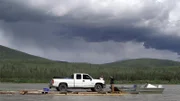 Joey Zuray (R), his sister, Kate Zuray (C), and cousin Zeb (L), transporting a truck down the Yukon River on a hand-built raft during the summer solstice. Joey is sitting, driving the boat.