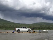 Joey Zuray (R), his sister, Kate Zuray (C), and cousin Zeb (L), transporting a truck down the Yukon River on a hand-built raft during the summer solstice. Joey is sitting, driving the boat.