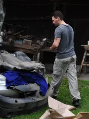 Ein Mann vor seinem Schuppen mit einem Auto mit offener Motorhaube und Werkzeug auf dem Tisch.